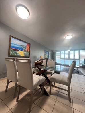 Un coin repas avec une table en verre, quatre chaises, des luminaires au plafond et un tableau accroché au mur.
