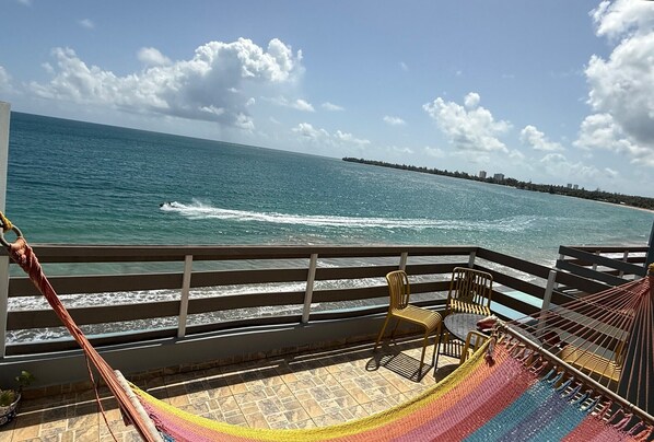 Uma rede e cadeiras numa varanda com vista para um corpo de água e uma costa distante.