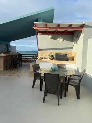 Um pátio externo com mesa e cadeiras de vidro, churrasqueira e vista para o oceano.