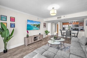 Ein modernes Wohnzimmer mit einem grauen Sofa, einem Couchtisch und einem Fernseher. Es gibt eine Küchenzeile mit einer Mikrowelle und einem Esstisch.