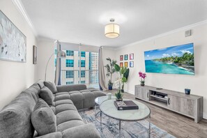 Ein modernes Wohnzimmer mit einem Ecksofa, einem Glastisch, einer TV-Bank und einem großen Fenster mit Blick auf ein Gebäude.