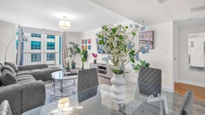 Un salon moderne avec une table basse en verre, des canapés gris et une grande fenêtre donnant sur des immeubles.