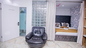 A modern bedroom with a large bed, a black leather chair, a glass shower, and a geometric-patterned wall. - Hidden Haven Apartment  (Lagos)