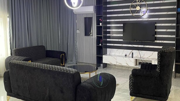 A modern living room with a black sofa, a matching armchair, a TV on a marble wall, and a ceiling with green and yellow lighting. - Hidden Haven Apartment  (Lagos)