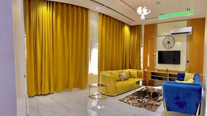 A modern living room with a yellow sofa, a blue sofa, a coffee table, and a TV mounted on the wall. - Hidden Haven Apartment  (Lagos)