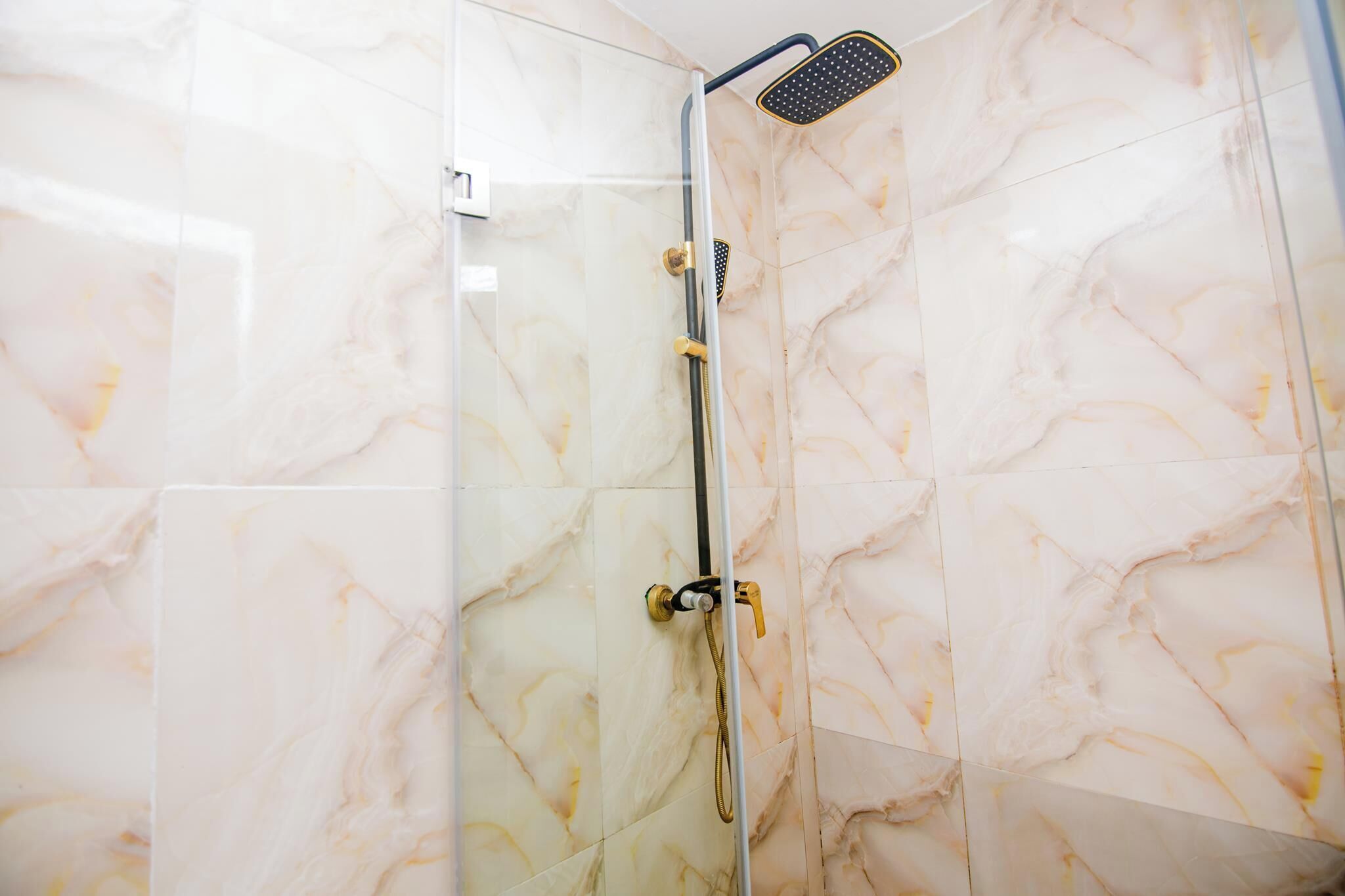A glass-enclosed shower with a handheld showerhead and a vertical rain showerhead, set against marble-patterned walls.