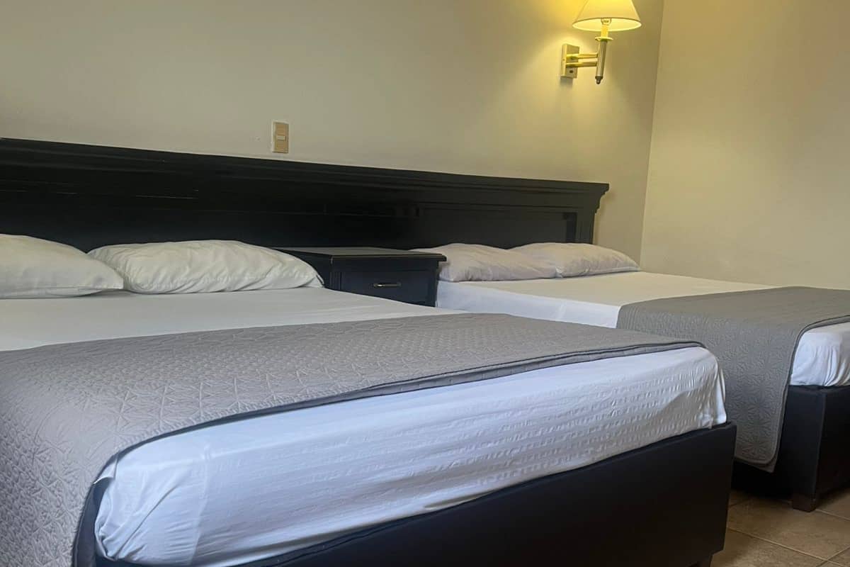 Une chambre d’hôtel avec deux lits, une tête de lit sombre, une lampe fixée au mur et un sol carrelé.