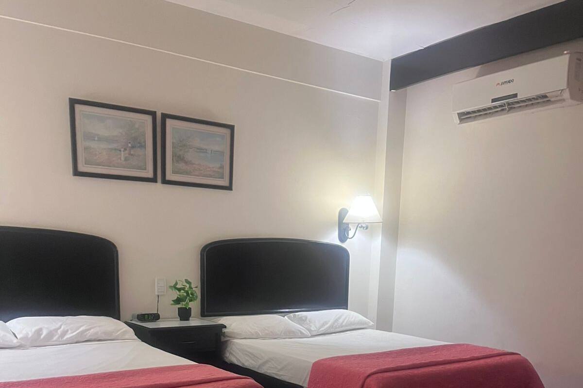 Une chambre d’hôtel avec deux lits, une table de chevet, une lampe, une horloge et un climatiseur fixé au mur.
