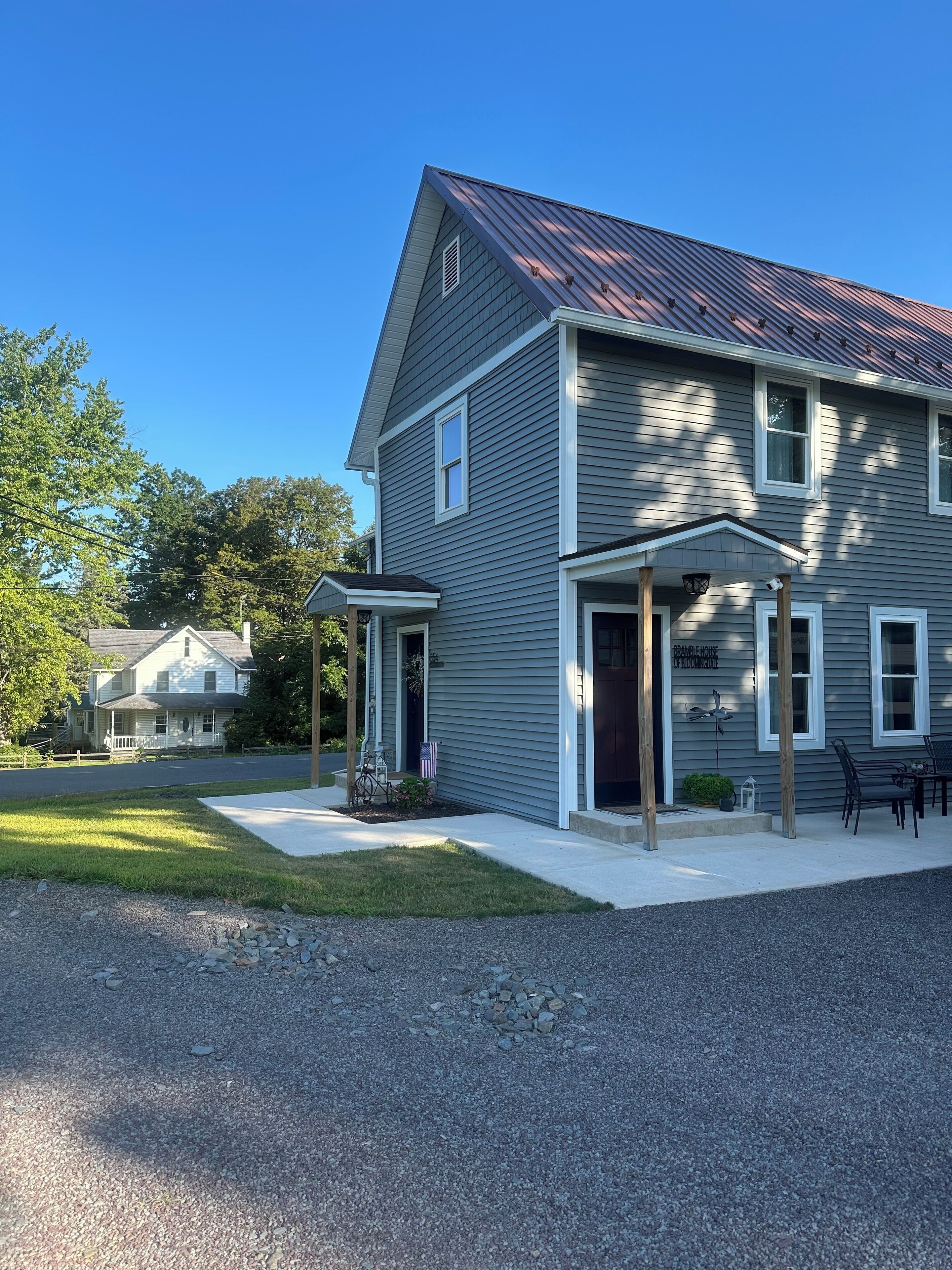 A two-story house with a covered porch, a front door, and a small garden area with a bench and an American flag.