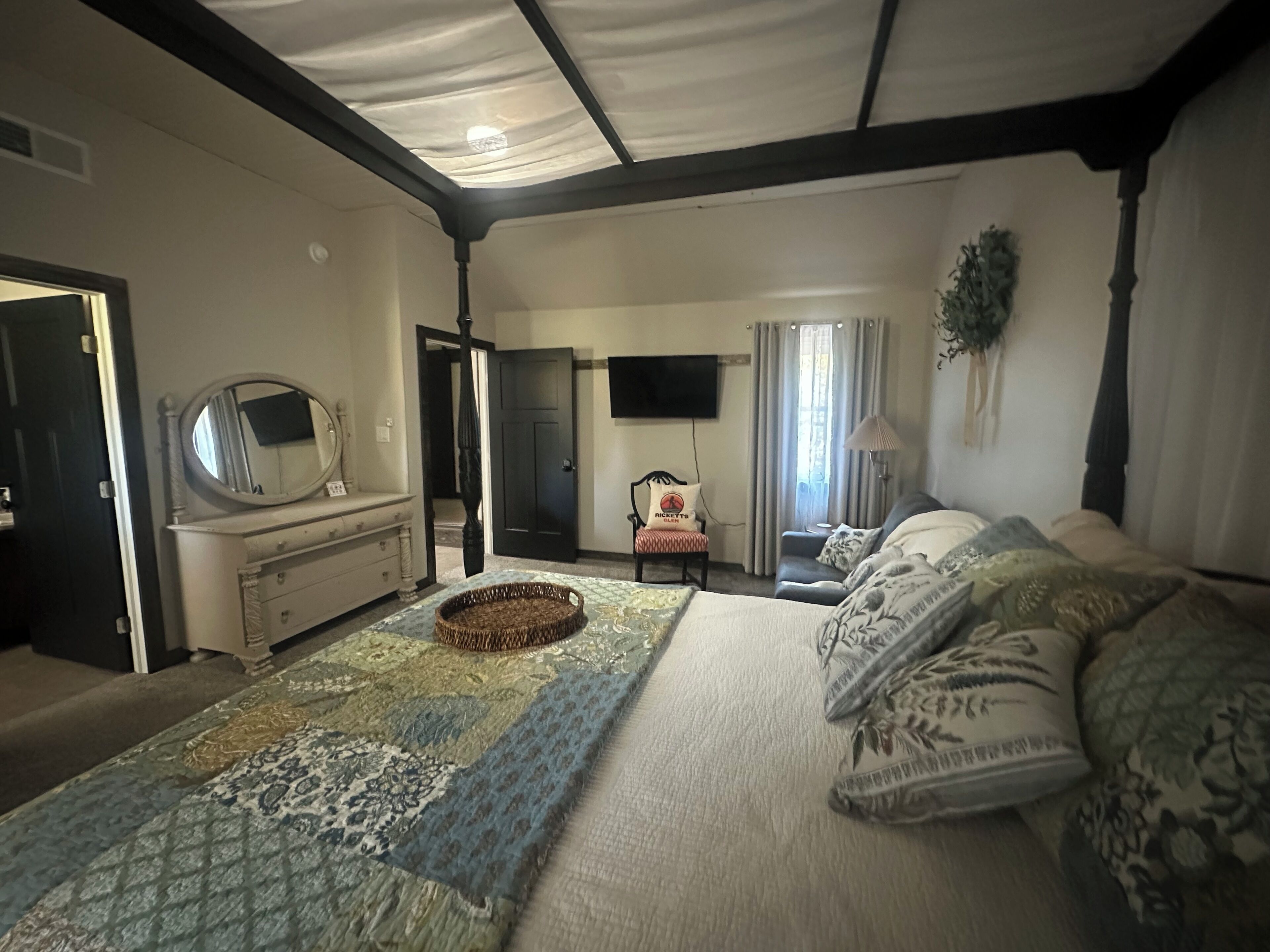 Quarto com uma cama grande, penteadeira com espelho, televisão na parede e porta que dá para outro cômodo.