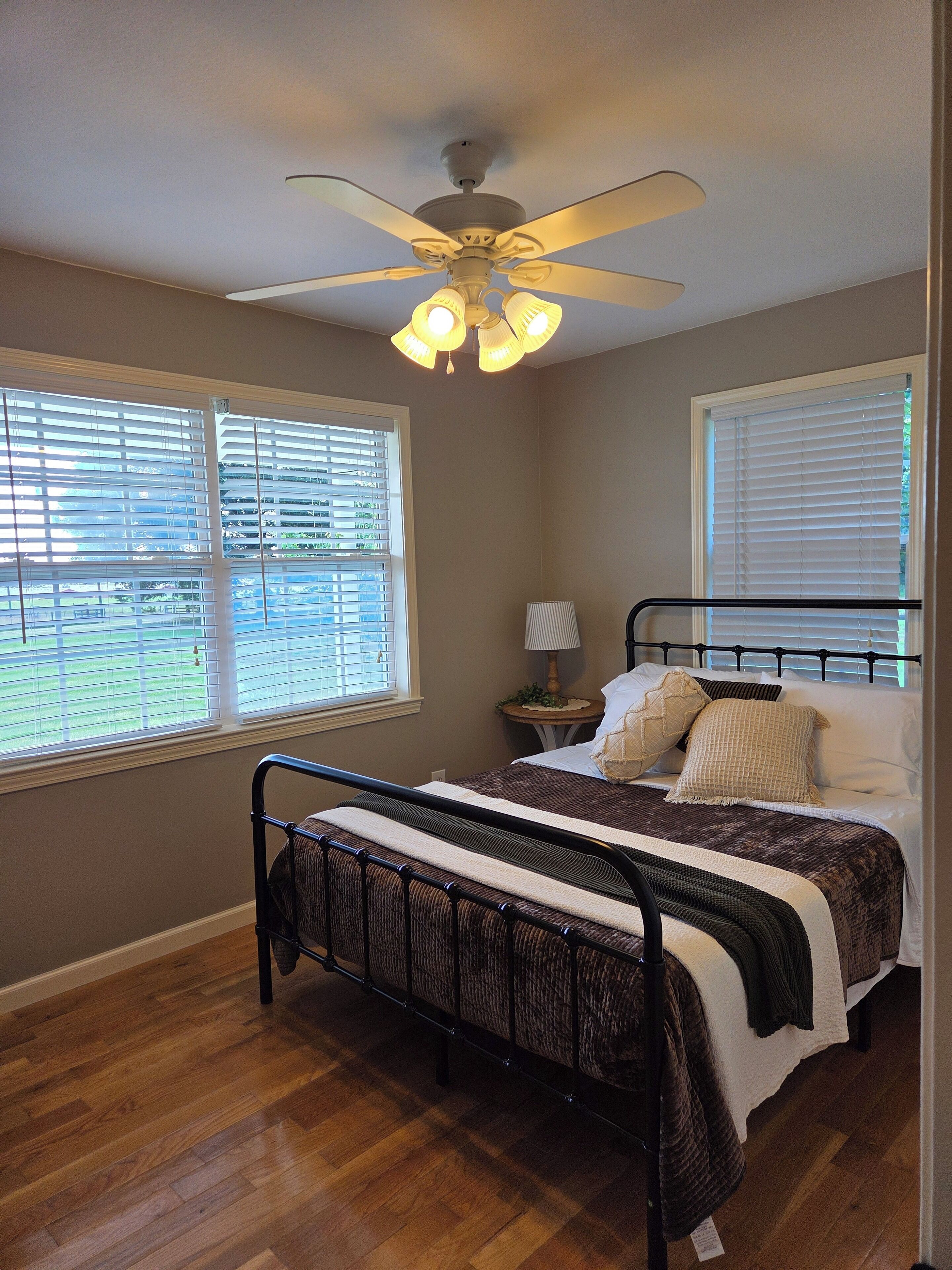 A bedroom with a bed, a ceiling fan, a nightstand, and two windows with blinds.