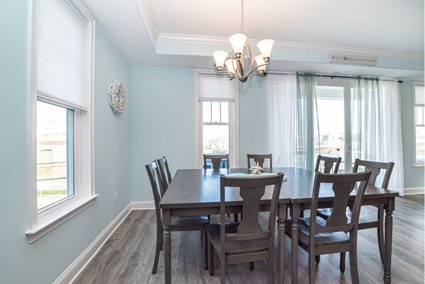 A dining room with a wooden table and chairs, a chandelier, and a view of the outdoors through large windows.