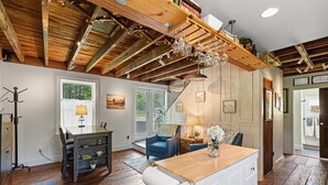 Un intérieur rustique avec des poutres apparentes, un coin repas avec une table et des chaises, une cuisine dotée d’un plan de travail en bois et un couloir orné de tableaux encadrés.