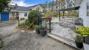 Une terrasse en bois avec des sièges d’extérieur, entourée de plantes en pot et d’un chemin de gravier menant à une maison avec une porte bleue.