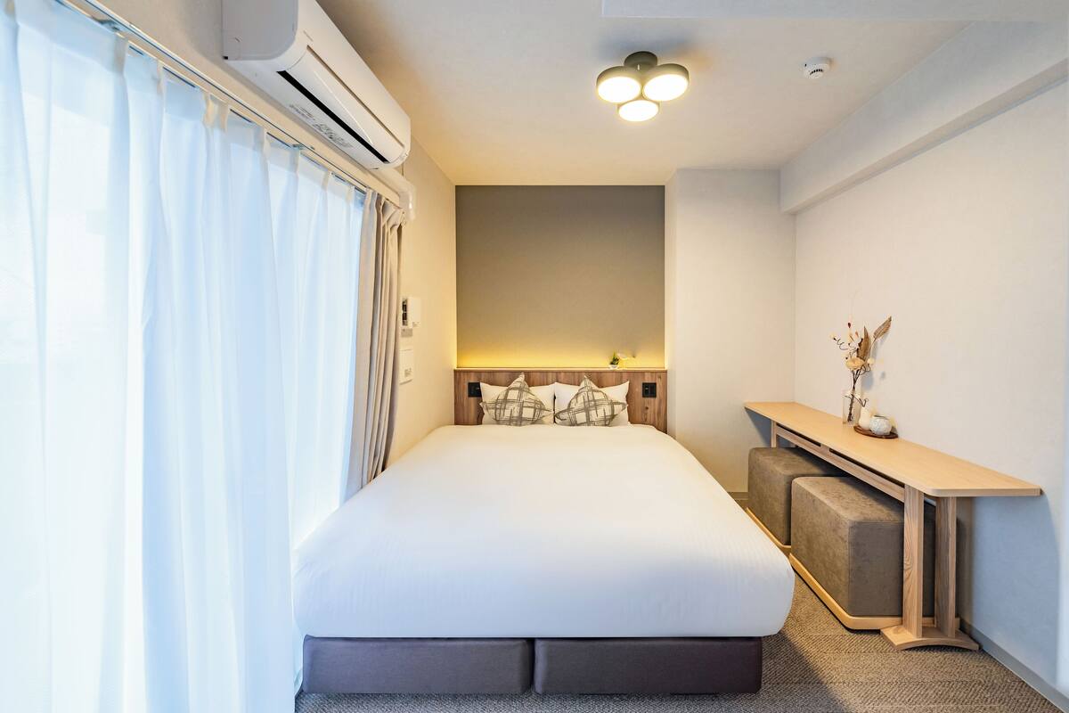 A hotel room with a large bed, a bedside table, a desk, and an air conditioning unit.