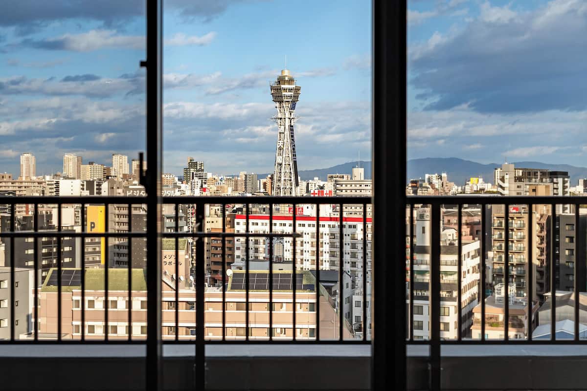 A view from a window with a metal railing, overlooking a cityscape with buildings and a tower.