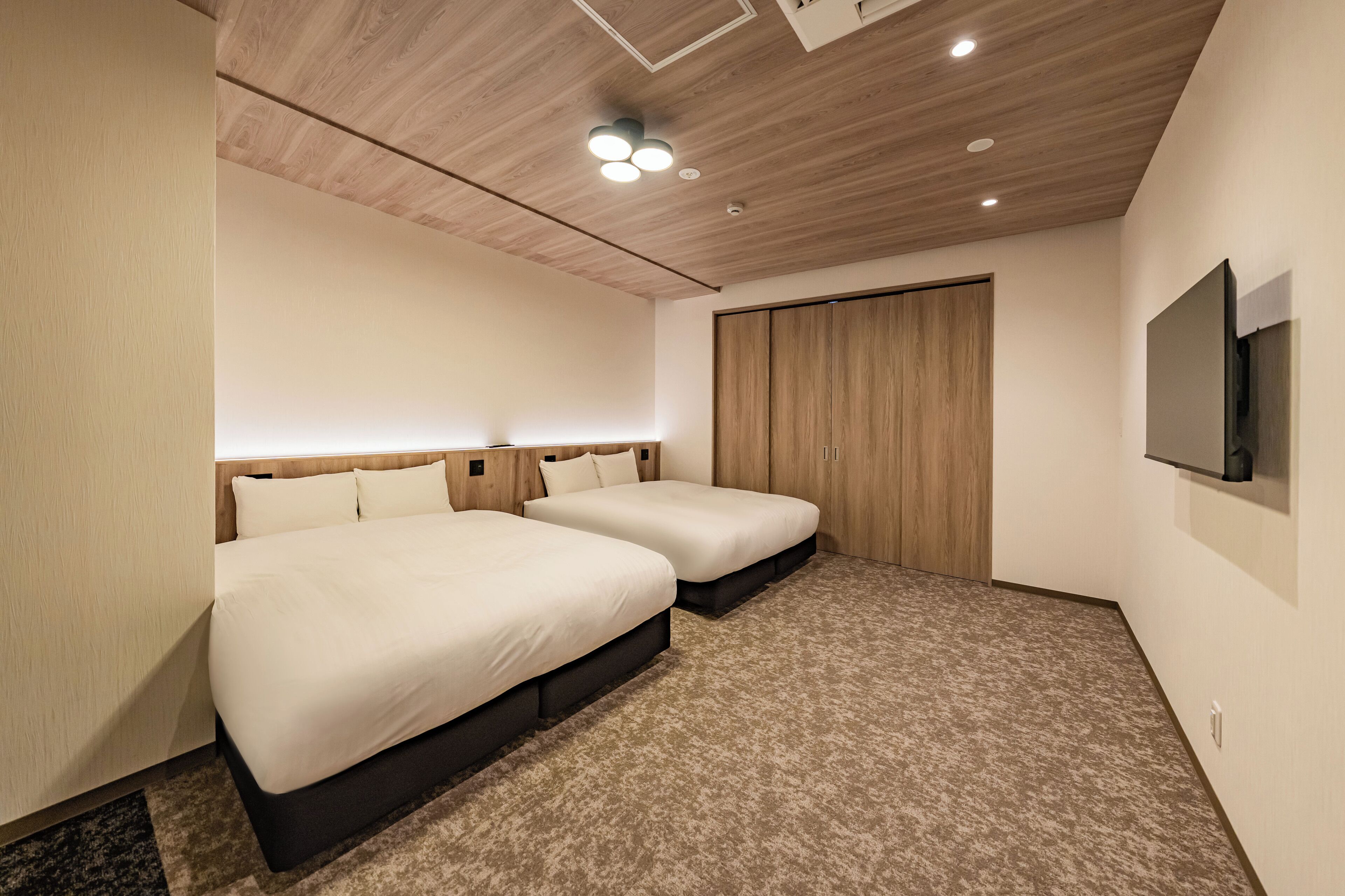 A hotel room with two beds, a wooden ceiling, a carpeted floor, a wardrobe, and a TV mounted on the wall.