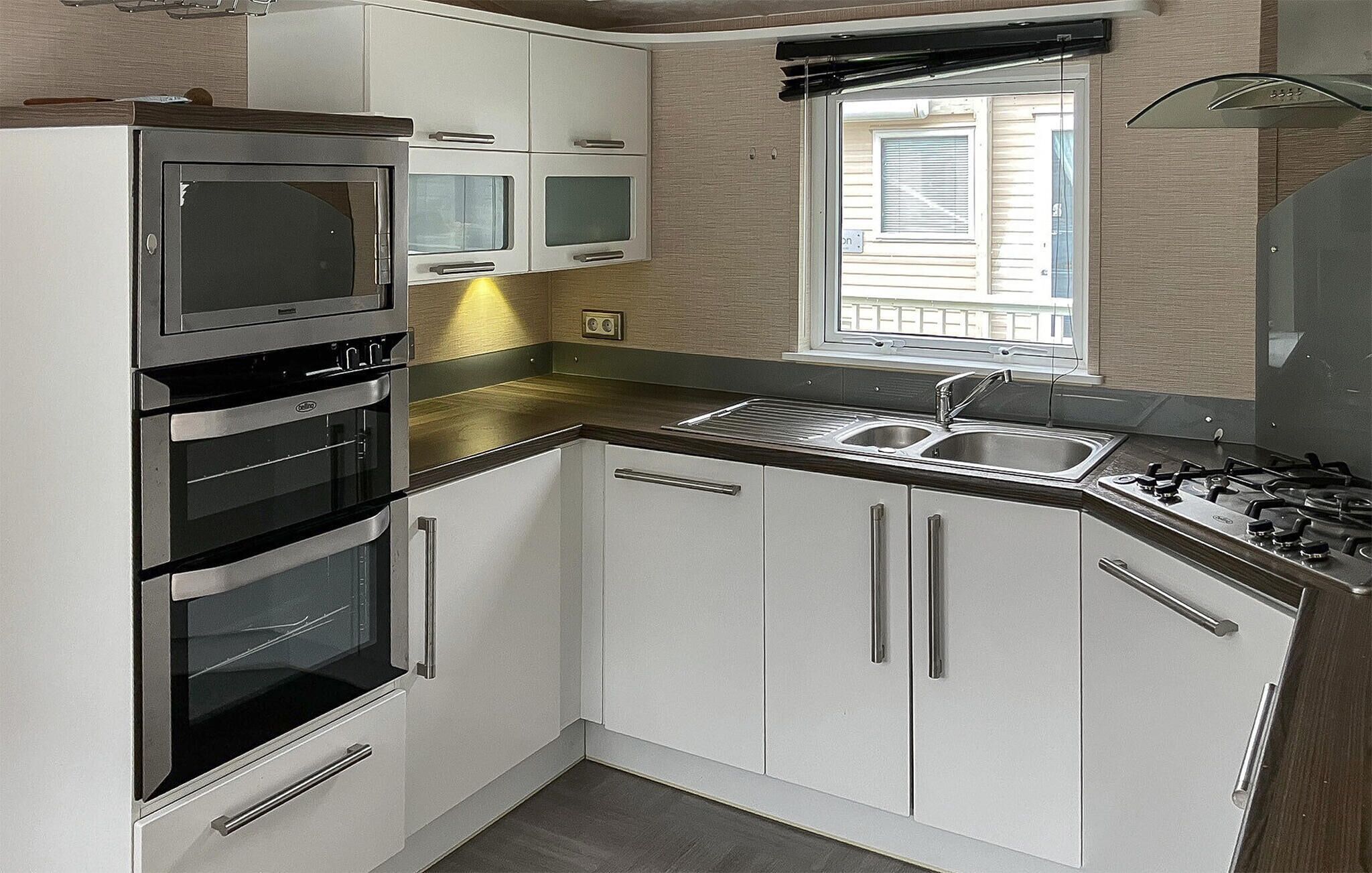 Una cocina compacta con armarios blancos, horno integrado, lavamanos y una ventana con persianas.