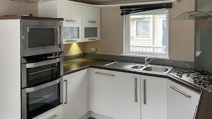 Una cocina compacta con armarios blancos, horno integrado, lavamanos y una ventana con persianas.