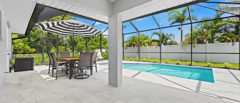 A covered patio with a pool, outdoor furniture, and a view of palm trees.