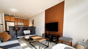 Una sala de estar moderna con sofá, una mesa de centro y televisor, una zona de cocina con armarios y microondas, y una pared de acento de madera.