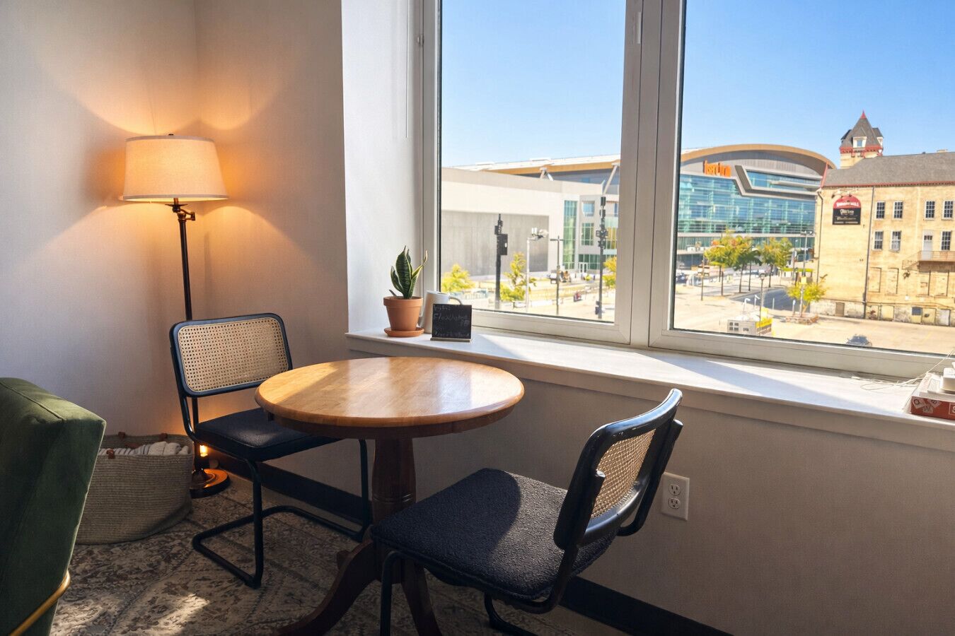 Een kamer met een ronde houten tafel, twee stoelen, een lamp, een potplant en een raam met uitzicht op een modern en een traditioneel gebouw.