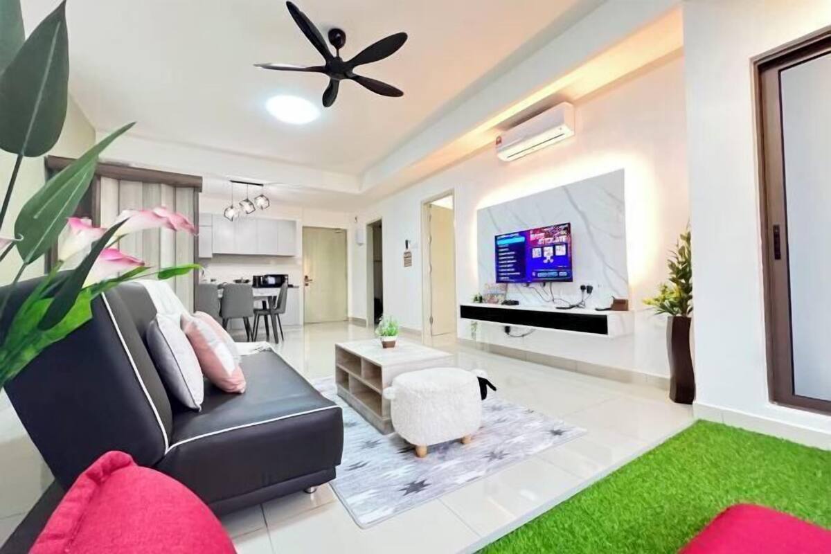 A modern living room with a black sofa, a white ottoman, a ceiling fan, a flat-screen TV, and a kitchen in the background.
