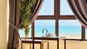 A room with a large window, a wooden table with a lamp, and a view of the beach.