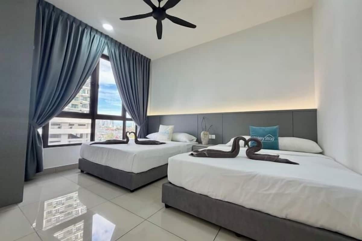 A hotel room with two beds, a ceiling fan, a large window with curtains, and a view of buildings outside.