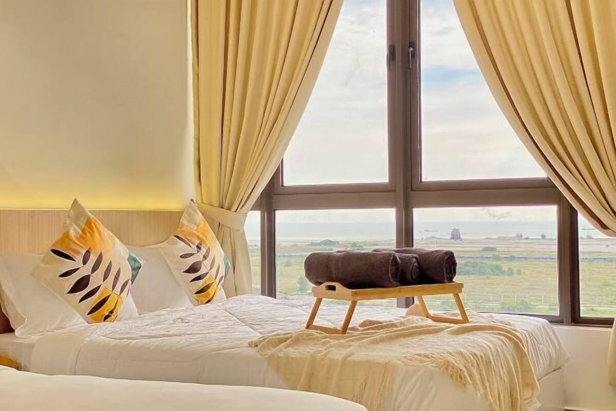 A bed with a view of the countryside through large windows, a bedside table with a towel, and decorative pillows.