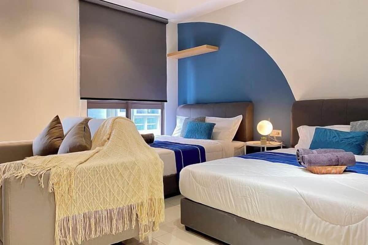 A hotel room with two beds, a sofa, a large window with blinds, and a nightstand with a lamp.
