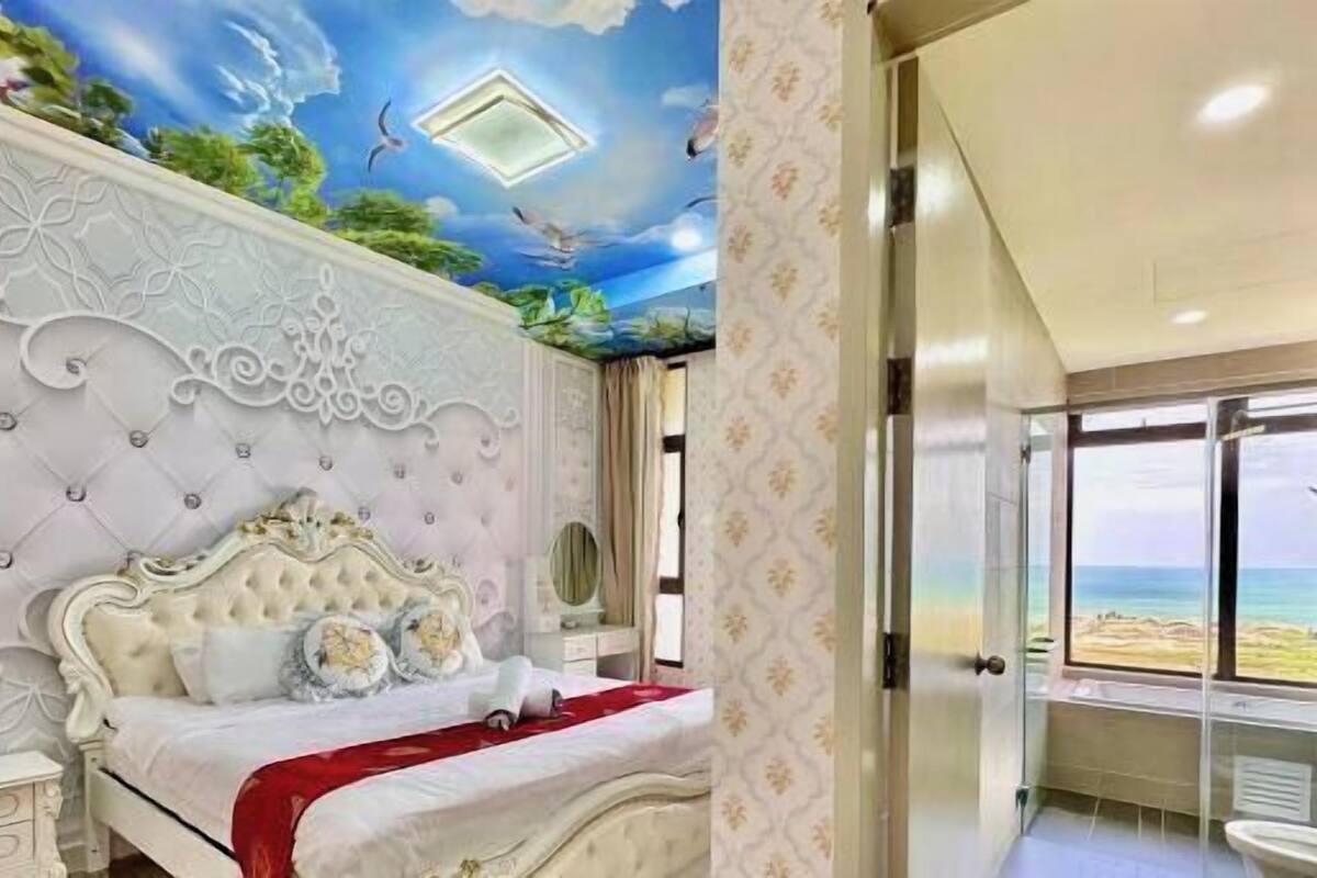 A bedroom with a bed, a bathroom with a shower, and a view of the beach.