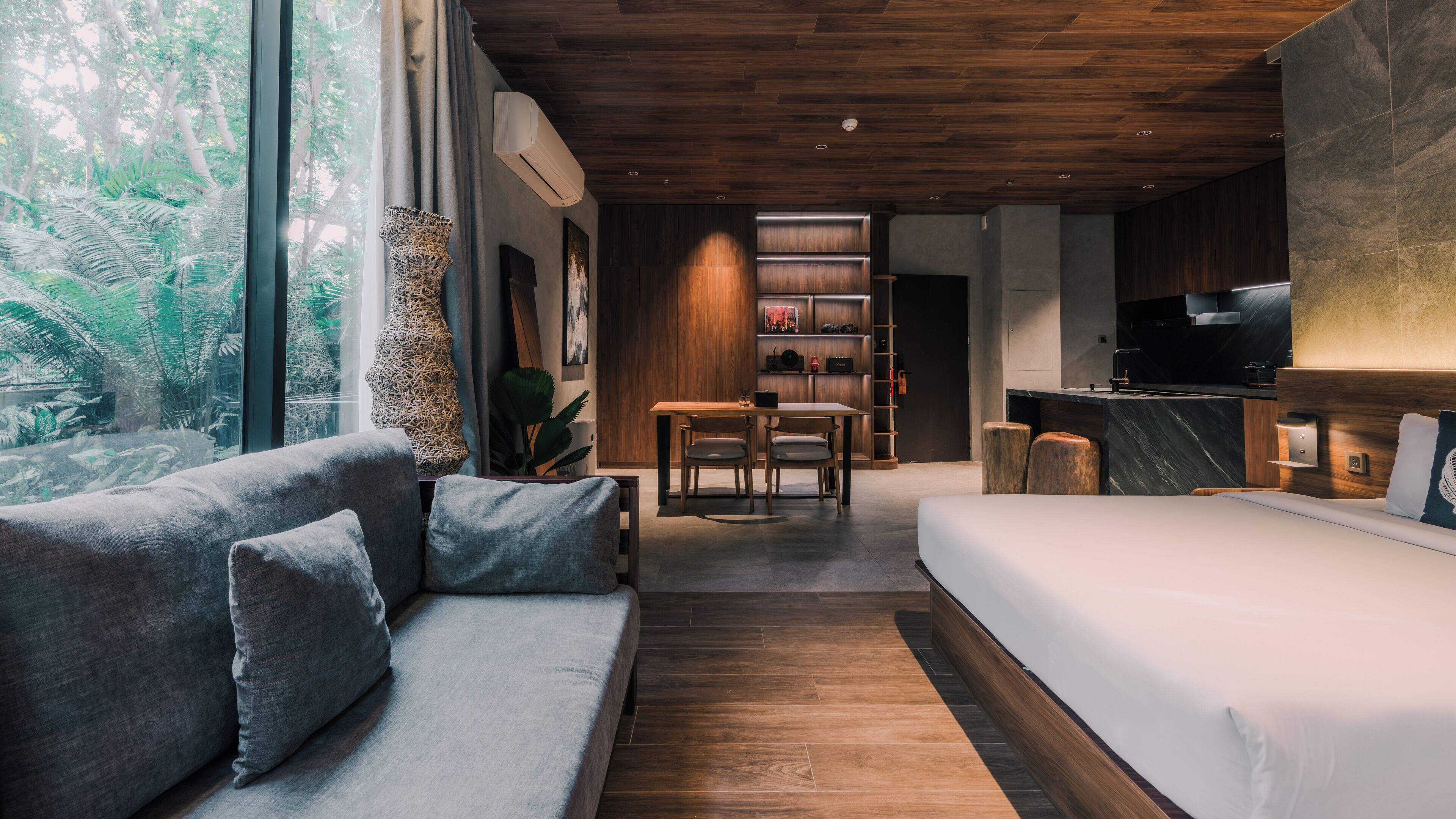 A modern hotel room with a wooden ceiling, a large window with a view of greenery, a sofa, a bed, a dining table with chairs, and a kitchen area.