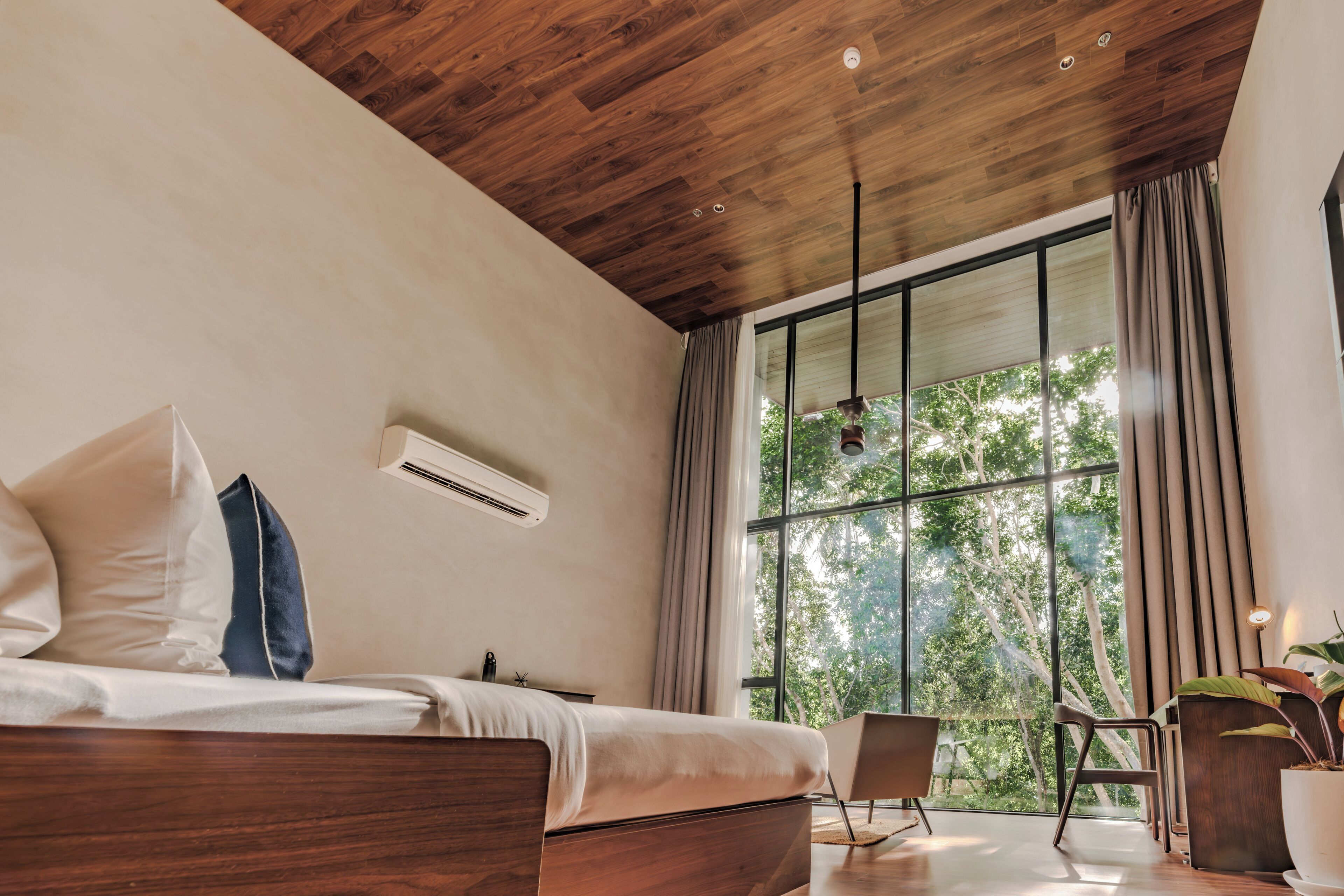 A modern bedroom with a wooden ceiling, a large window with curtains, a bed with pillows, a chair, and a potted plant.