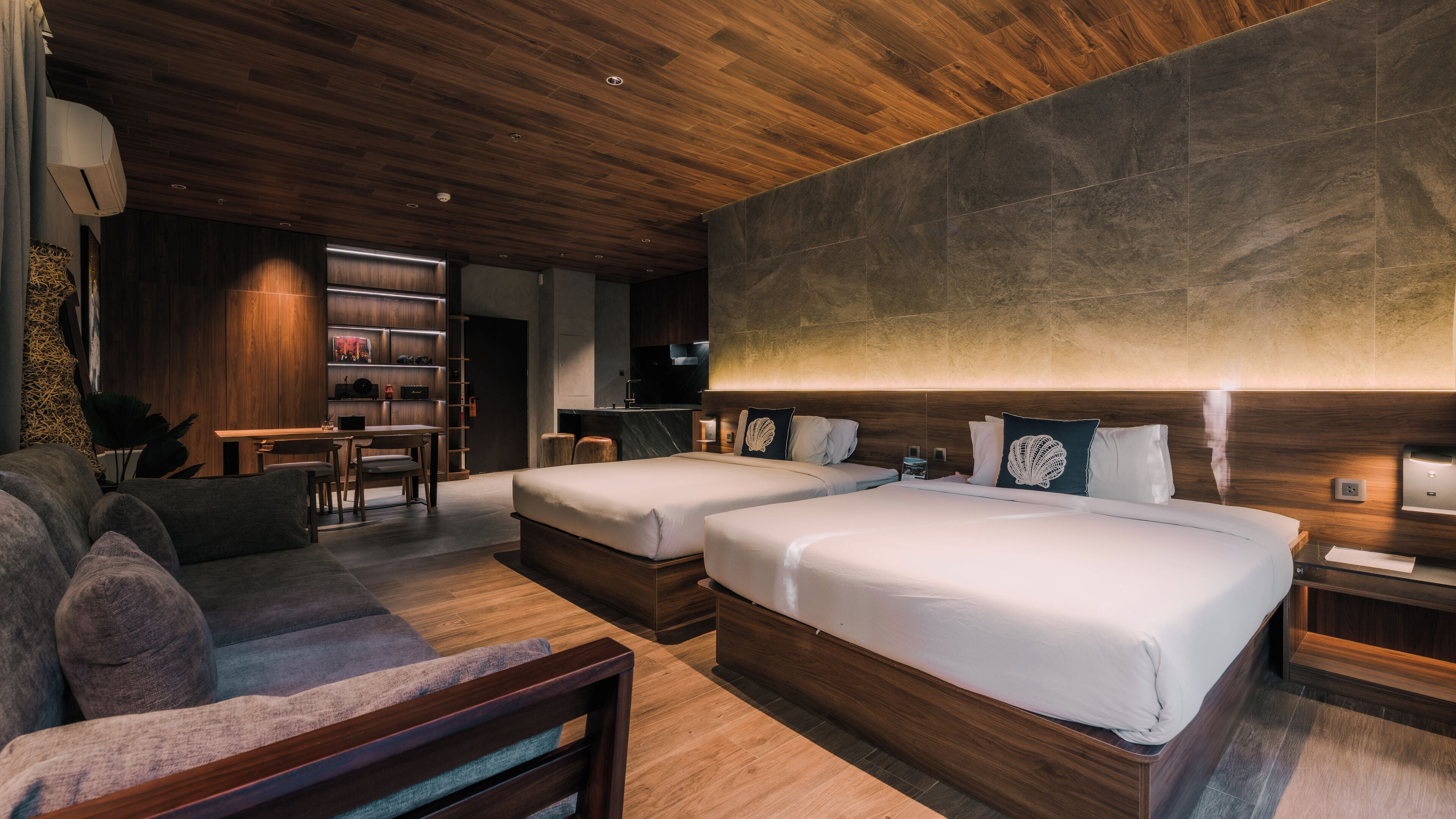 A modern hotel room with two beds, a sofa, a desk, and a kitchen area.