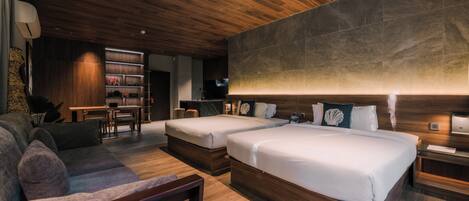 A modern hotel room with two beds, a sofa, a desk, and a kitchen area.