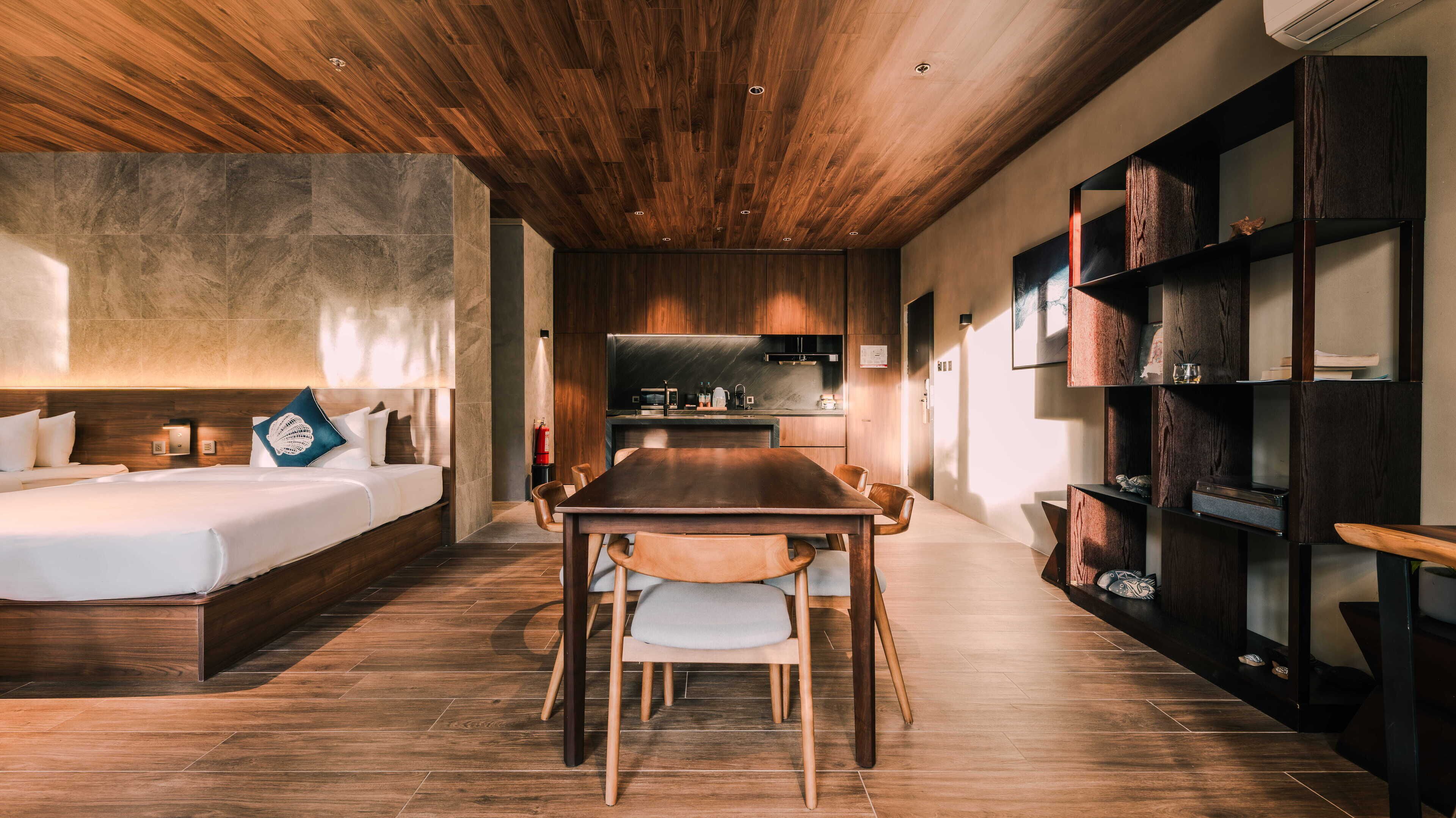 Una habitación de hotel moderna con techo de madera, una cama grande, una mesa de comedor con sillas, una zona de cocina y estanterías empotradas de madera.