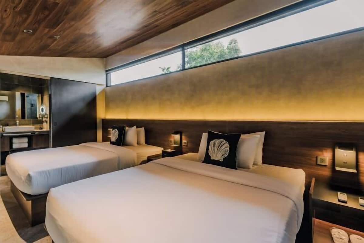Een hotelkamer met twee bedden, een groot raam, een houten plafond en een badkamer met spiegel en wastafel.