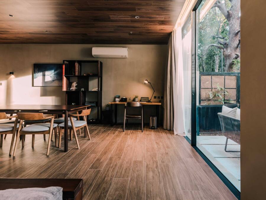 Una habitación moderna con techo de madera, una mesa de comedor con sillas, un escritorio con silla, una lámpara de pie y vistas a zonas verdes al exterior.