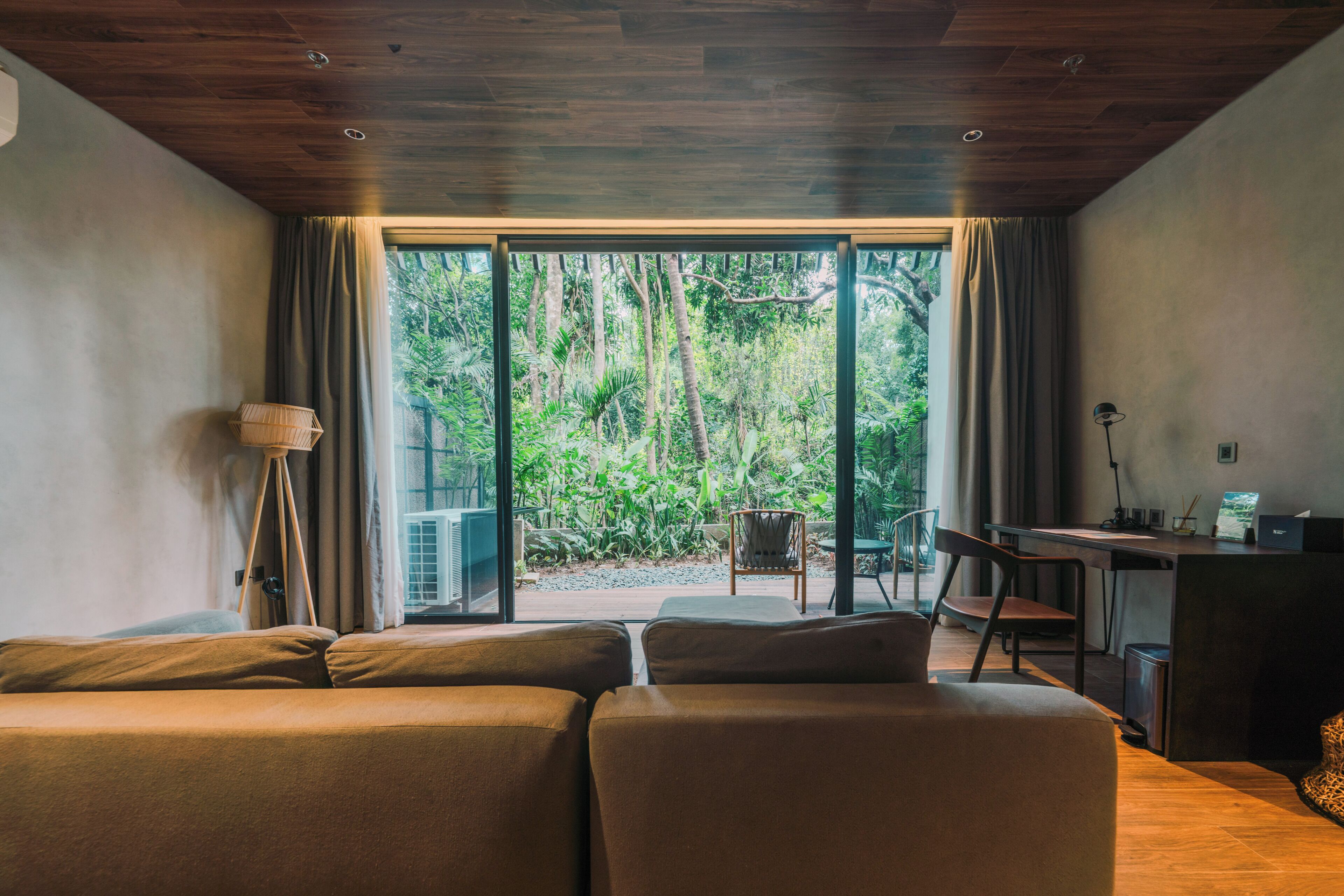 Una habitación con techo de madera, un sofá, un escritorio y vistas a un bosque a través de un ventanal.