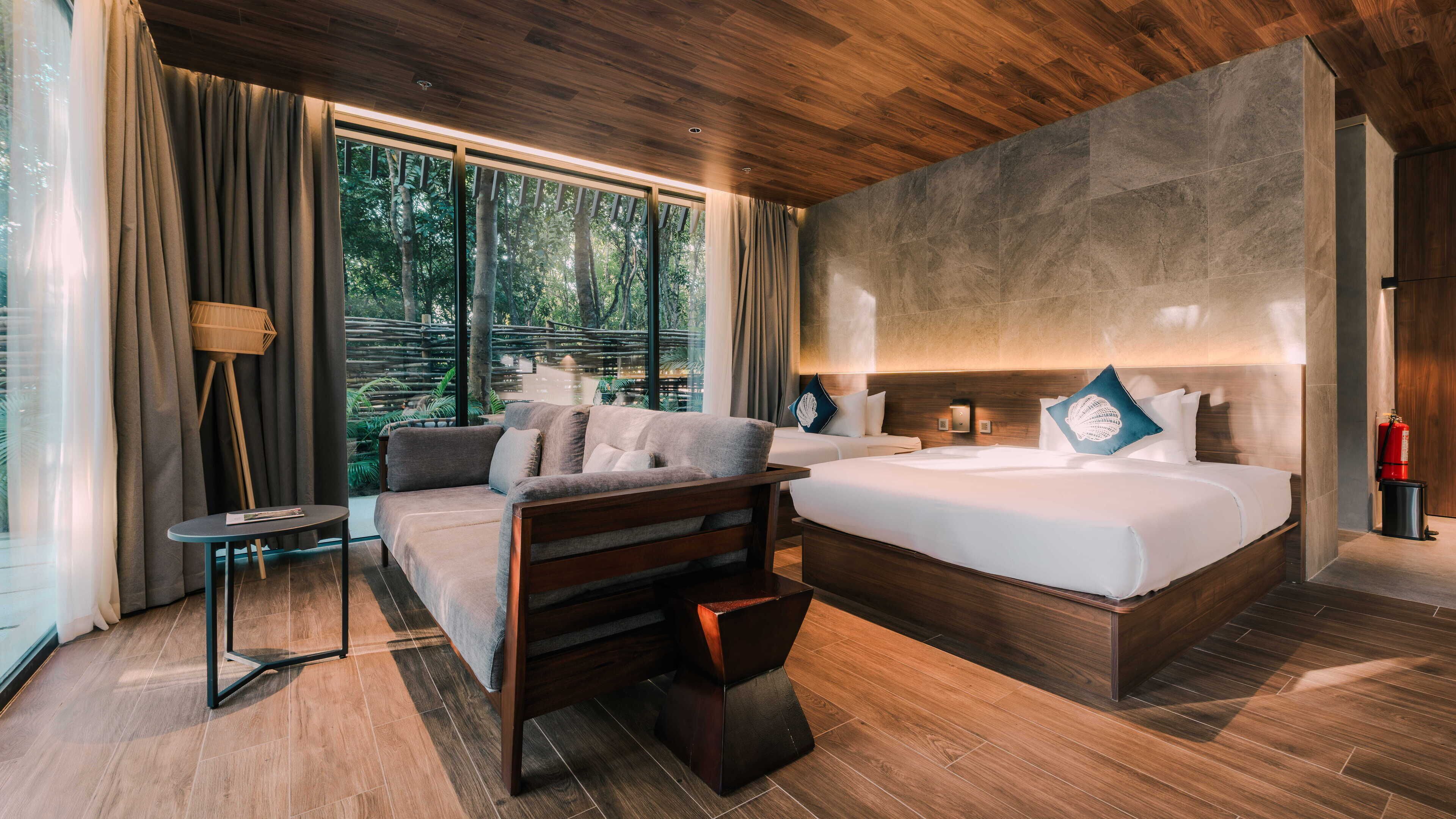 Ein modernes Hotelzimmer mit einem großen Bett, einer Couch, einem kleinen Tisch und Blick auf einen Wald durch große Fenster.