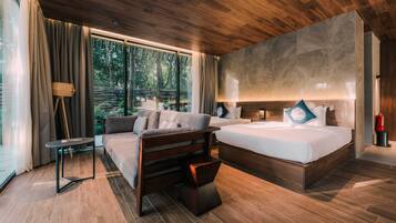 Ein modernes Hotelzimmer mit einem großen Bett, einer Couch, einem kleinen Tisch und Blick auf einen Wald durch große Fenster.