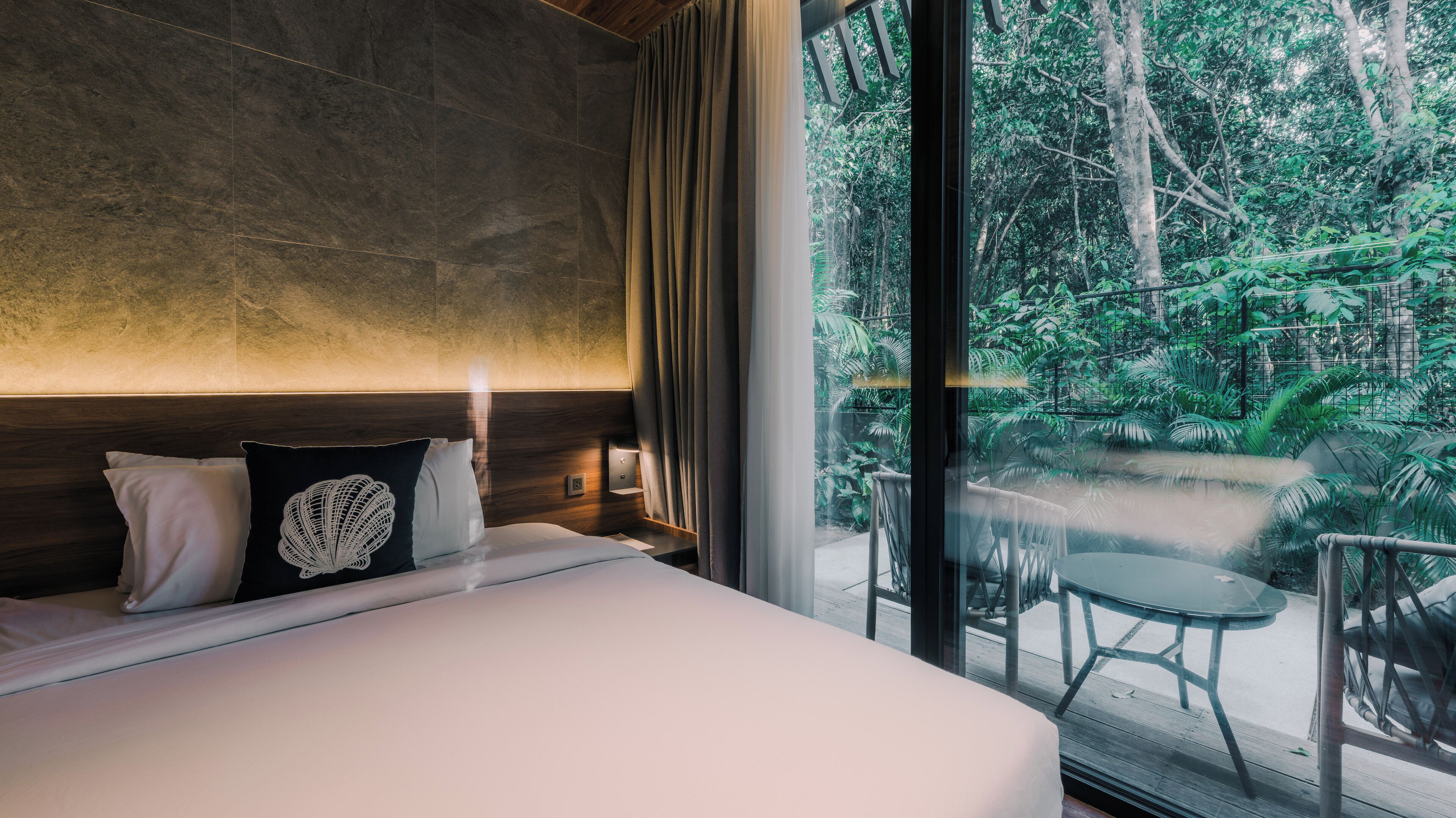 Quarto com uma cama grande, uma mesa de cabeceira, janela com vista para uma floresta e varanda com mesa e cadeiras.