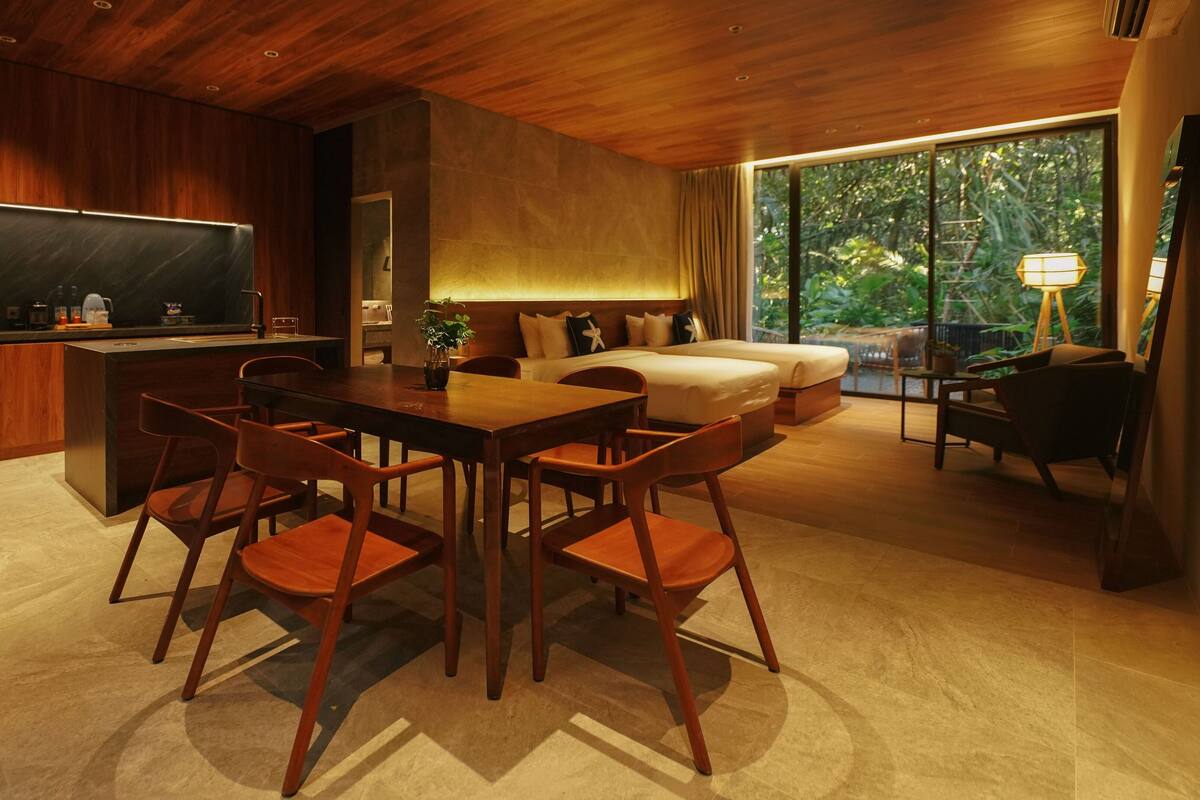 Een moderne hotelkamer met een houten plafond, een zithoek met een tafel en stoelen, een bank, een bed en een groot raam met uitzicht op groen.