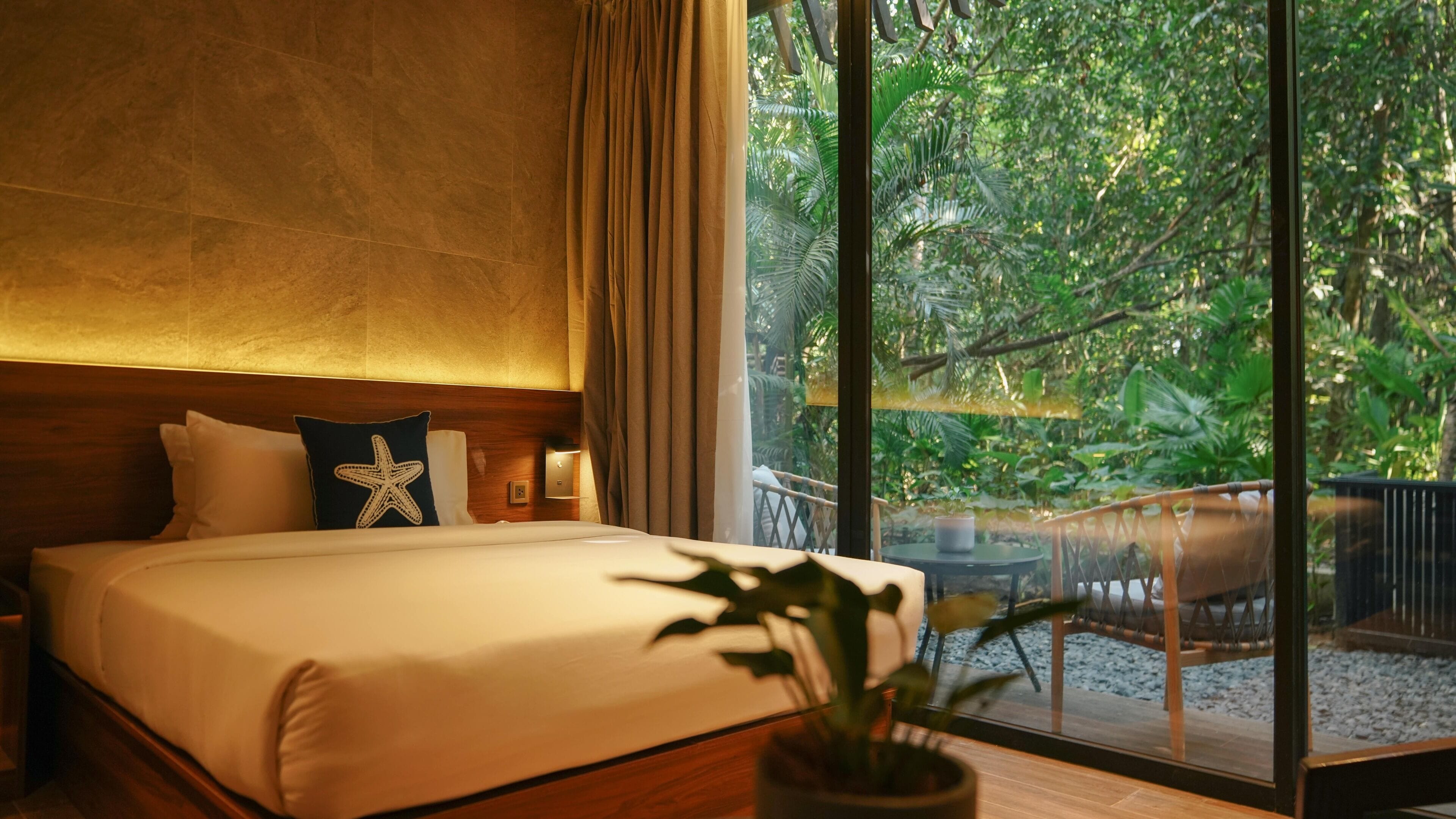 Une chambre à coucher avec un lit, une table de chevet, un coussin en forme d’étoile, une lampe de chevet et une vue sur un balcon agrémenté de plantes.