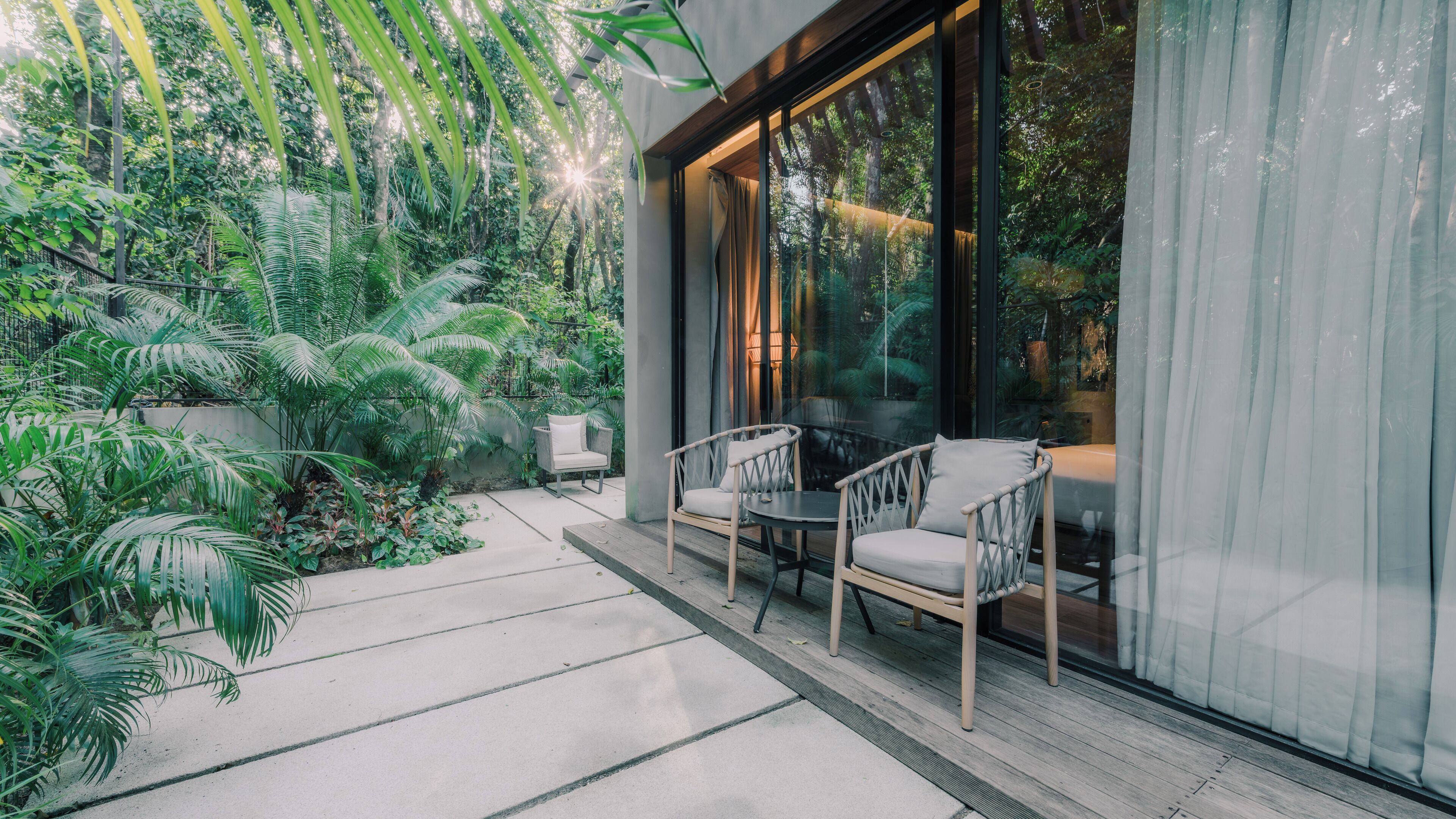 A modern outdoor seating area with chairs and a table, surrounded by lush greenery and a glass wall.