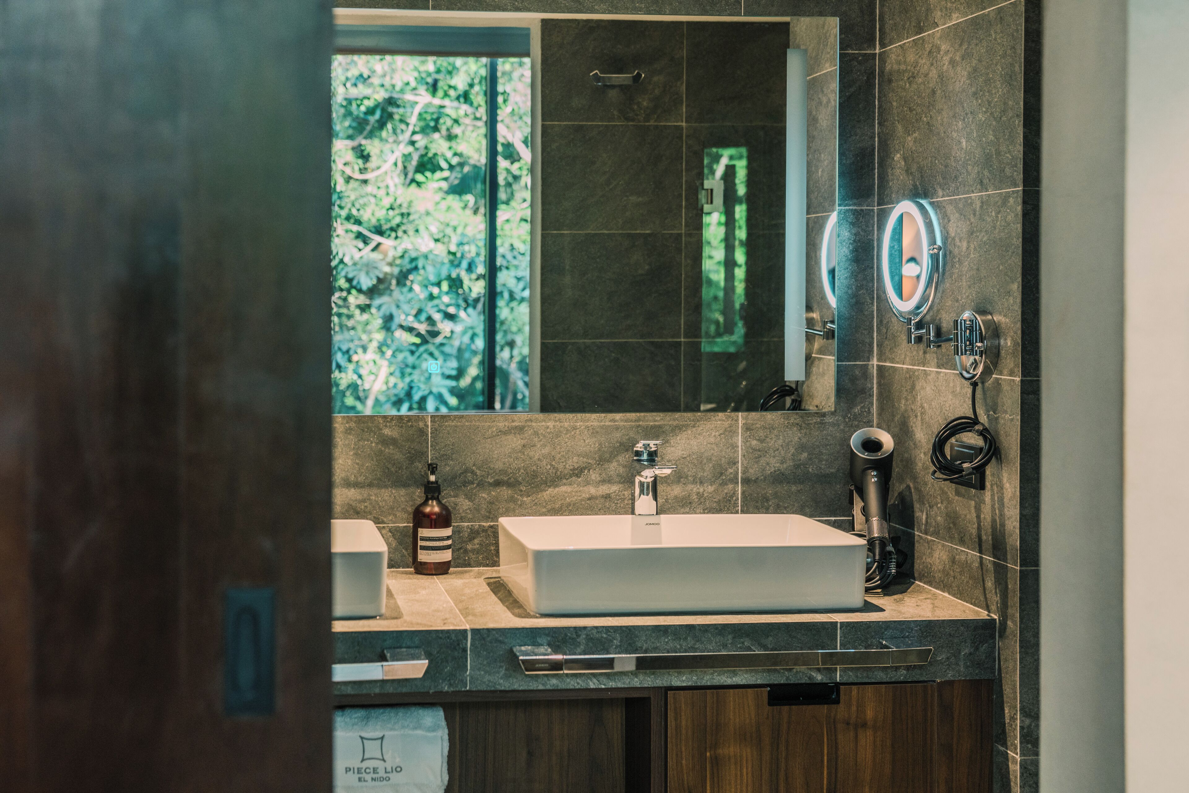 Un baño moderno con un espejo grande, un lavamanos rectangular y una ventana con vistas a la vegetación.