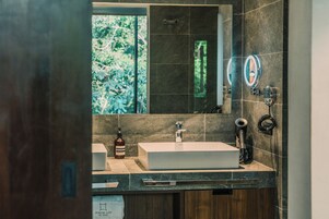 Un baño moderno con un espejo grande, un lavamanos rectangular y una ventana con vistas a la vegetación.