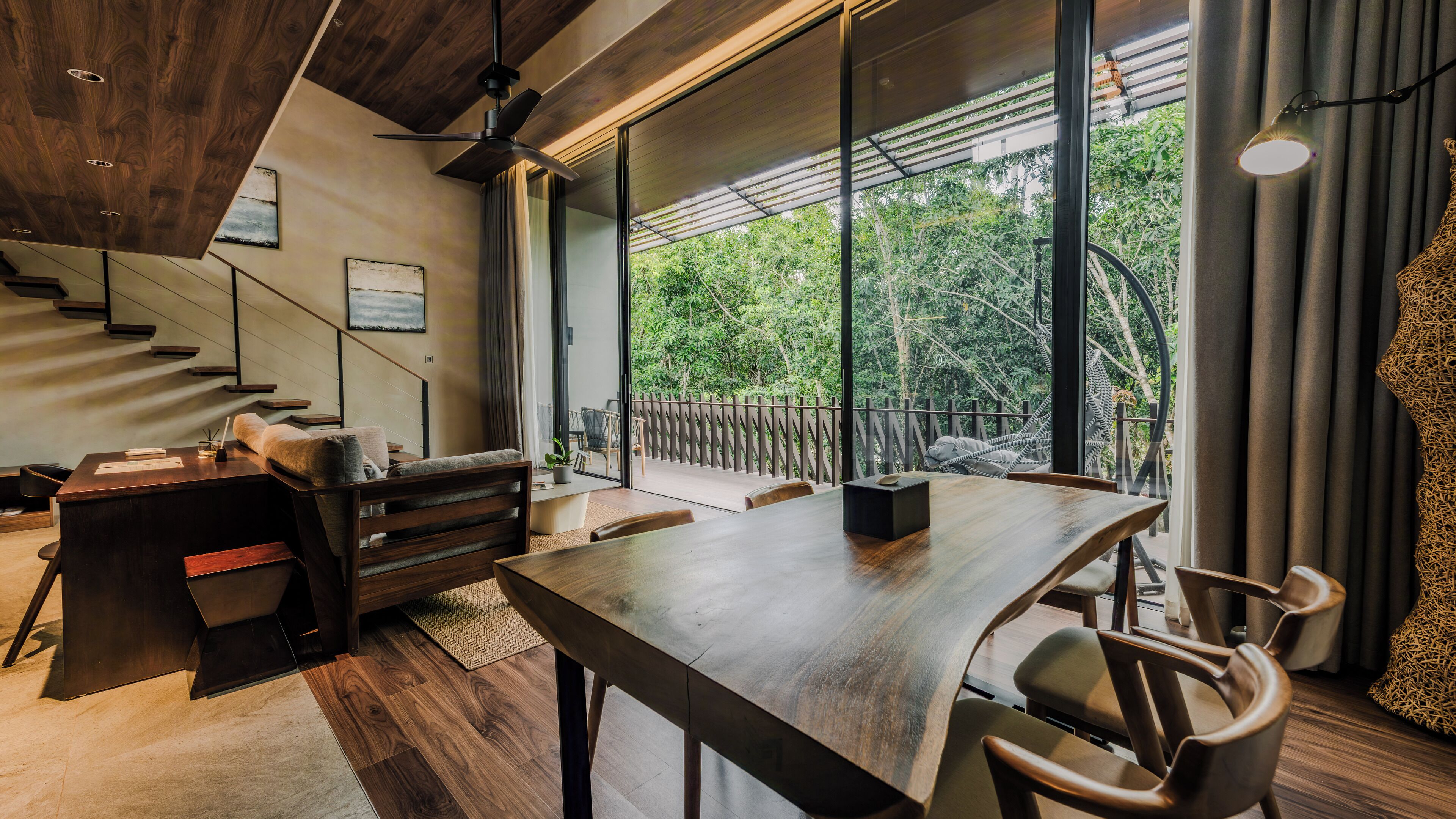 A modern interior with a wooden dining table, chairs, a sofa, a staircase, and large windows offering a view of greenery.