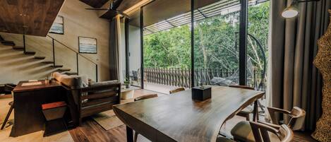 A modern interior with a wooden dining table, chairs, a sofa, a staircase, and large windows offering a view of greenery.